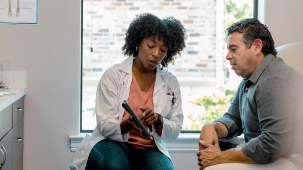 Physician sits with man in grey sweater and point to a tablet.