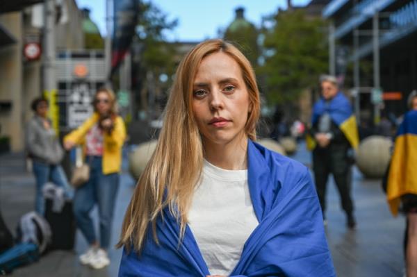 Ukrainian refugee Olesia, who has long blo<em></em>nde hair and is wearing a white t-shirth with her country's blue and yellow flag around her shoulders, at a rally for Ukraine in Sydney's Martin Place