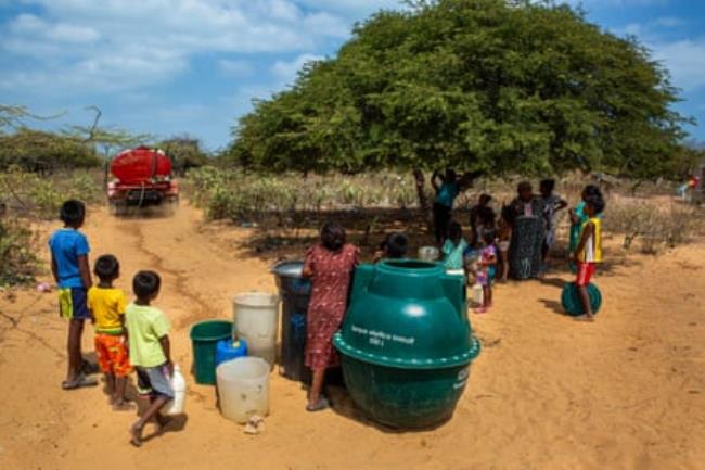 A group of adults and children surrounded by water co<em></em>ntainers watch as a truck drives off down a dirt road