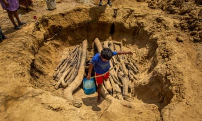A young boy carries a co<em></em>ntainer of water away from a hole in the sandy ground. The hole has logs of varying sizes over it forming two small platforms with a gap in the middle for drawing water.