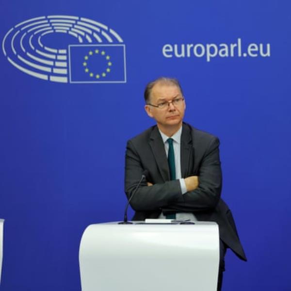 Philippe Lamberts standing with his arms folded behind a podium, against a bright blue wall with the EU flag on it and the text 