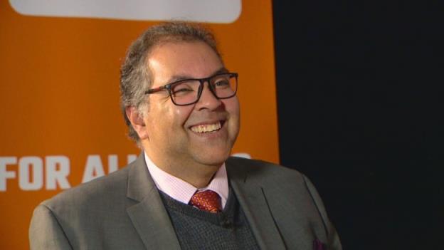 A man wearing a suit smiles in front of a backdro<em></em>p.