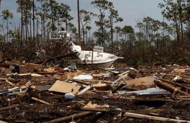 A white boat amid completely leveled ground covered in wood and dotted with palm trees, beneath a bright, gray sky.