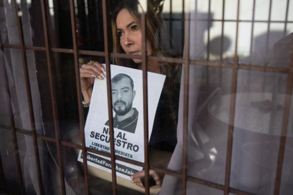 A woman holds up a missing person's poster with her brother's face on it, as she stares through a window protected with me<em></em>tal bars.