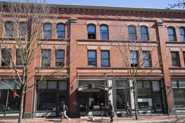 A person looks out the window as others walk by an affordable housing building run by Central City Concern, a Portland-ba<em></em>sed homeless services nonprofit, on Friday, March 15, 2024, in Portland, Ore. The no<em></em>nprofit leases more than 1,000 single room occupancy units, both subsidized and not, to people who are co<em></em>nsidered extremely low-income. It helps people struggling to access housing due to things like eviction histories and poor credit scores. (AP Photo/Jenny Kane)