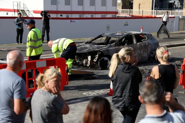 o<em></em>nlookers gather near a burnt and destroyed car.