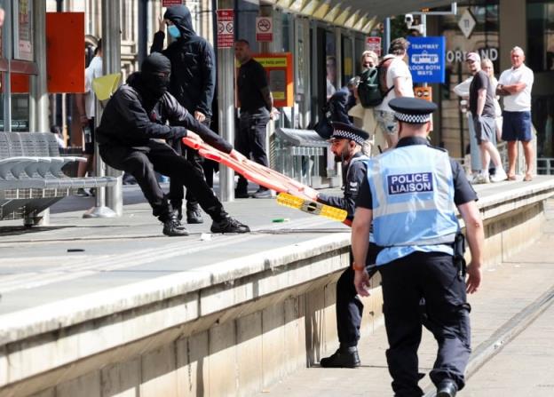 A masked protestor and a police officer struggle for possession of a plastic barrier.