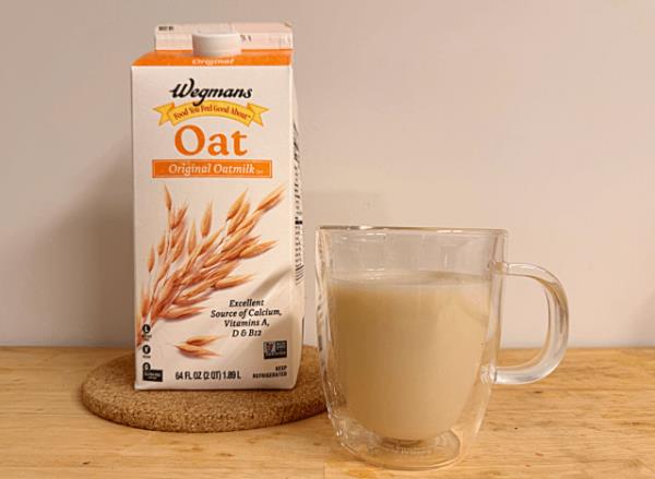 a co<em></em>ntainer of wegman's oat milk next to a glass mug filled with coffee and oat milk.
