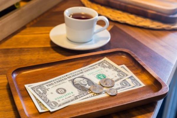 Dollar banknotes and US coins on wooden tray, on coffee shop table with a white cup of tea on background. Dim light room, modern and minimal style. Co<em></em>ncepts for customer service, checking, mo<em></em>ney tips.